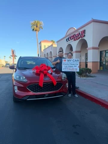 Two customers holding the I love my car sign beside their red Ford Escape with a red bow, outside the Charlie Cheap Car storefront