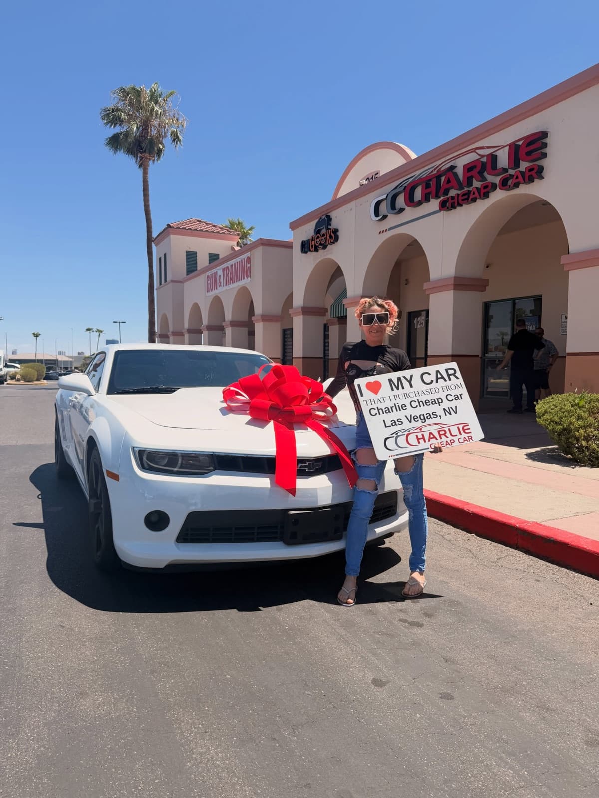A customer holding the I love my car sign beside a white Chevrolet Camaro with a big red bow, outside the Charlie Cheap Car storefront