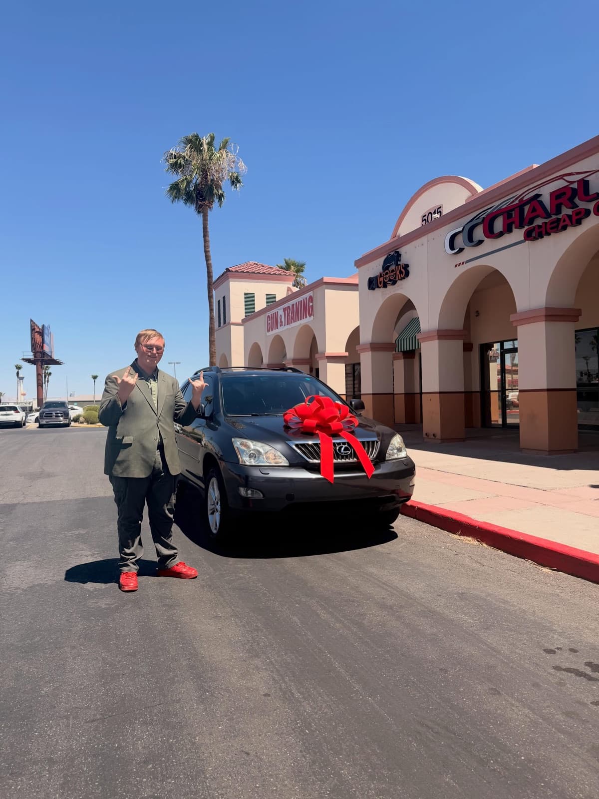 A happy customer giving a thumbs up beside a black Lexus SUV with a red bow, outside the Charlie Cheap Car storefront