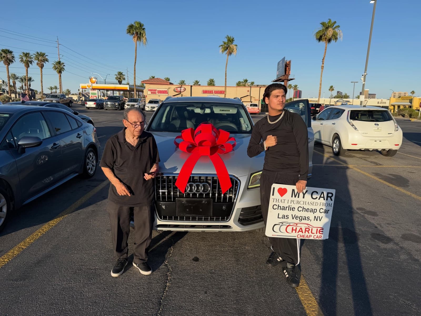 Two customers with the I love my car sign on either side of a white Audi Q5 with a big red bow, on the Charlie Cheap Car lot