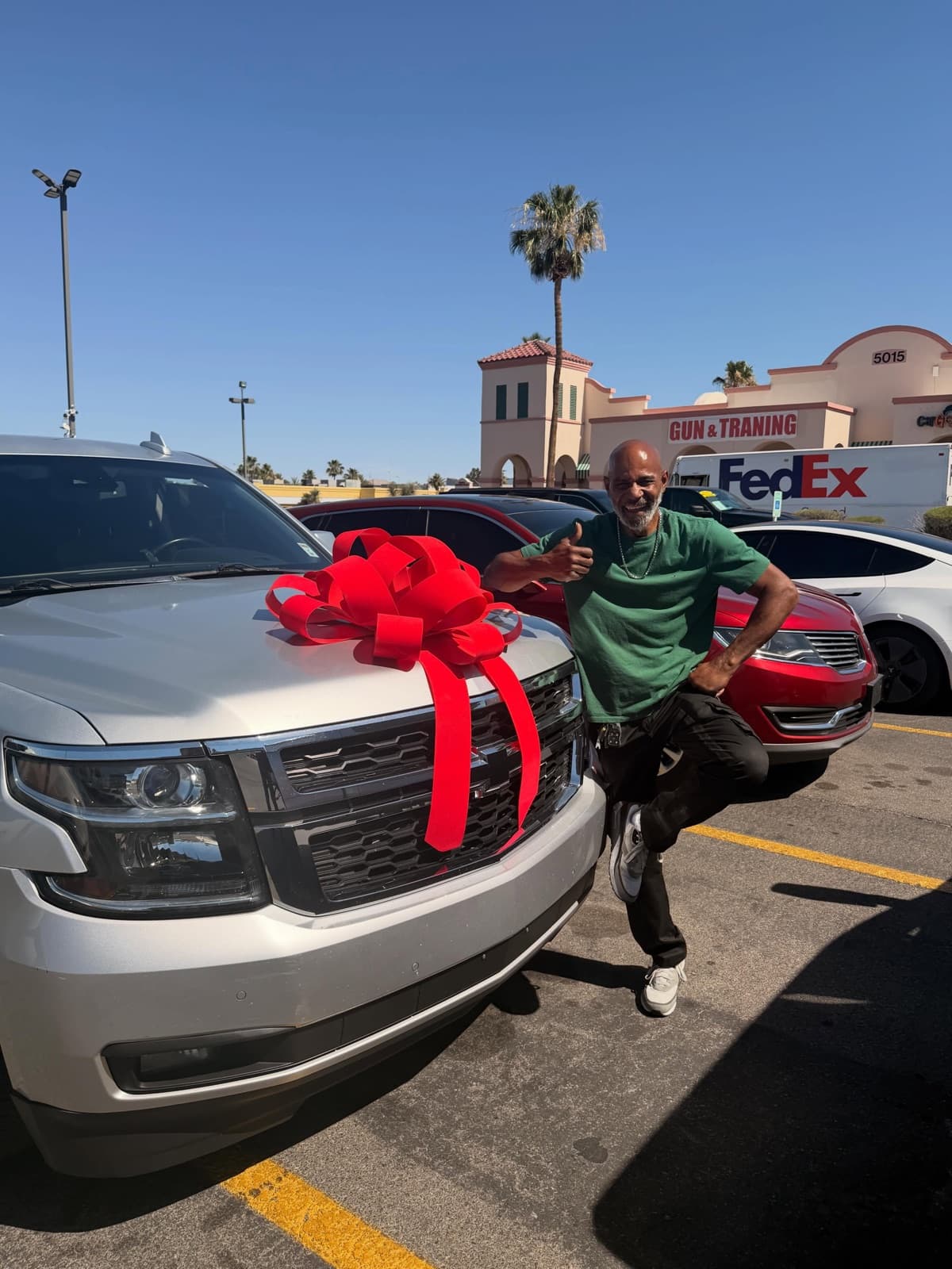 A smiling customer posing beside a silver Chevrolet Tahoe with a big red bow, on the Charlie Cheap Car lot